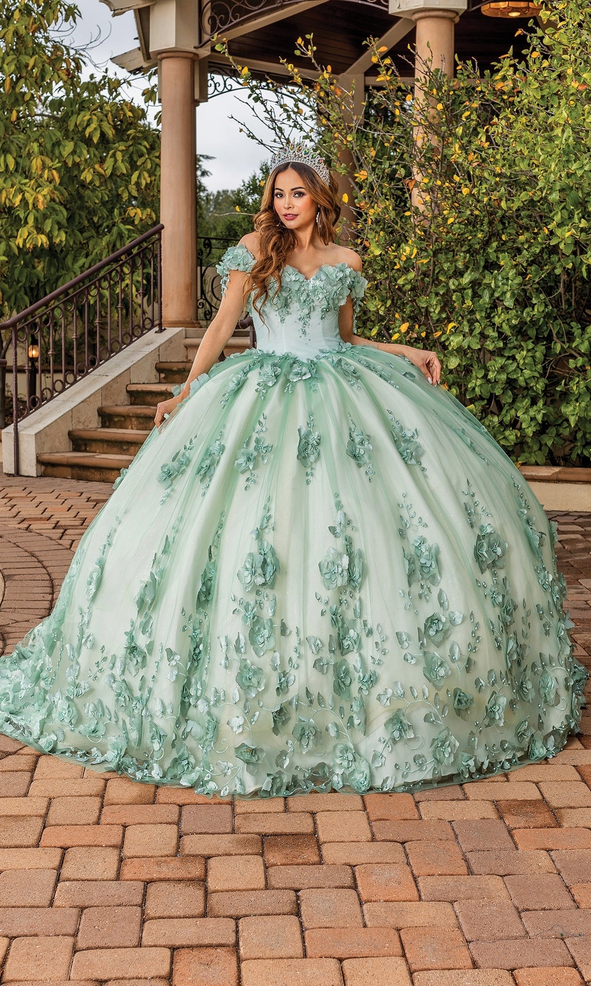 A woman stands outdoors on a stone path near greenery and a gazebo, wearing the Quinceanera Dress 1818 by Dancing Queen—a light green ball gown with floral appliqué, sweetheart neckline, and tiara.